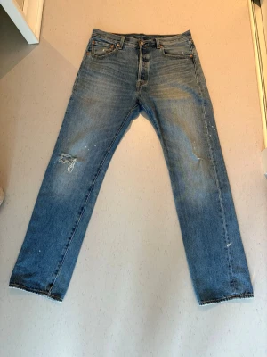 Levi's blå raka jeans med slitningar - Blå jeans från Levi's (501) med raka ben och klassisk femficksmodell. Jeansen har slitningar och hål på vänster lår samt lätt tvättad look. Tillverkade i denim med detaljer som nitar och läderpatch bak i midjan. W32 L32