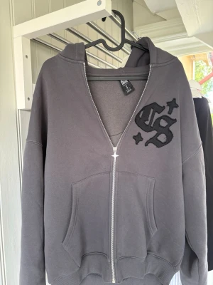 Grå zip hoodie  - Säljer en grå hoodie med dragkedja. Hoodien har stor svart broderad GS-logga med stjärnor både fram och bak, samt en rymlig huva och kängurufickor. Tillverkad i mjukt sweatshirtmaterial, perfekt för en chill och avslappnad stil.
