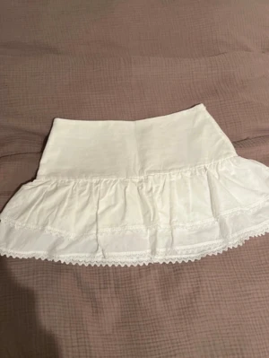 Vit volangkjol med spetsdetaljer - Supersöt vit kortkjol med volang och spetsdetaljer längs kanten. Kjolen har en feminin och luftig design som passar perfekt till sommaren. Tillverkad i mjukt bomullstyg för en bekväm känsla. Perfekt att matcha med en enkel topp eller stickad tröja.