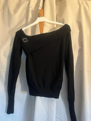 Svart off shoulder tröja med ribbad kant - Trendig svart off shoulder tröja med bred ribbad kant upptill och dekorativ metallspänne på ena axeln. Långärmad modell med åtsittande passform och ribbade muddar vid ärmslut och nederkant. Perfekt för en stilren och modern look.
