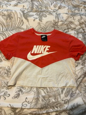 Röd och vit croppad Nike t-shirt - Säljer en croppad t-shirt från Nike i rött och vitt med stor logga framtill. T-shirten har korta ärmar och är tillverkad i mjuk bomull. Perfekt för en sportig och avslappnad stil.