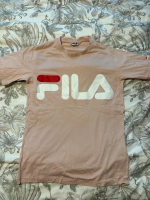 Beige FILA t-shirt med stor logga - Snygg beige t-shirt från FILA med stor vit och röd logga framtill. Klassisk rund halsringning och korta ärmar. Perfekt för en avslappnad och sportig stil.