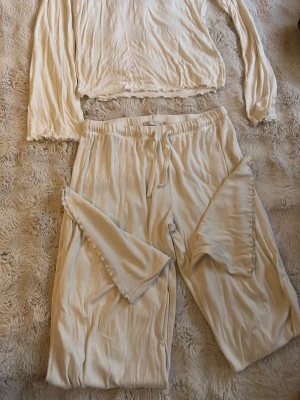 Beige loungewear-set med volangdetaljer - Mysigt loungewear-set i beige med långärmad topp och vida byxor. Setet har volangdetaljer vid ärmslut och benslut samt dragsko i midjan. Tillverkat i ett mjukt, glansigt material som känns skönt mot huden. Perfekt för hemmadagar eller chillkvällar.