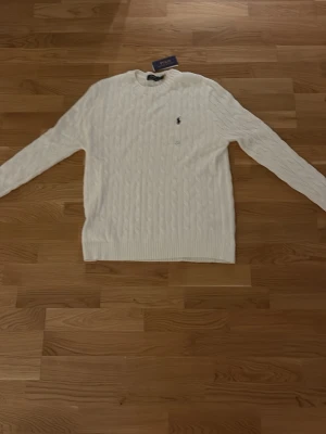 Vit kabelstickad tröja Polo Ralph Lauren - Kabelstickad vit tröja från Polo Ralph Lauren med rund halsringning och den klassiska lilla marinblå logotypen broderad på bröstet. Tröjan har ribbade muddar vid ärmslut och nederkant och är tillverkad i mjukt material.