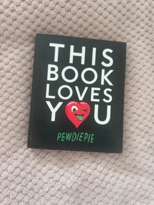 This Book Loves You av PewDiePie - Rolig och unik bok med svart omslag och stor vit text, samt en röd tecknad hjärta på framsidan. Perfekt för dig som gillar humor och memes, skriven av PewDiePie. Boken är i pocketformat och har en modern, lekfull design.