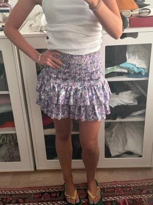 Ljuslila blommig volangkjol - Supersöt kort volangkjol i ljuslila med blommigt mönster. Kjolen har smock i midjan och dubbla volanger som ger en flowy känsla. Perfekt för sommarens alla tillfällen och lätt att matcha med en enkel topp.