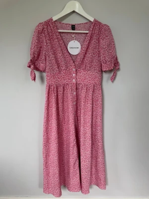Rosa blommig klänning från SHEIN, stl S - Söt rosa klänning från SHEIN med vitt blommönster, v-ringning och knappar framtill. Klänningen har korta ärmar med knyt och en luftig, avslappnad passform. Perfekt för varma dagar och somriga vibes. För kännedom så var det svårt att få med den äkta rosa färgen på bilderna. Rökfritt hem.