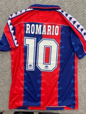 Barcelona Kappa Romario tröja - Retro Barcelona matchtröja från Kappa med Romario och nummer 10 på ryggen. Tröjan har klassiska blå och röda vertikala ränder, klubbmärke på bröstet och Kappa-loggor på ärmarna. Perfekt för dig som älskar fotboll och vintage stil.
