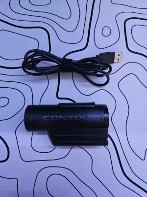 Contour kamera - Säljer en svart actionkamera från Contour med cylindrisk form och robust design i plast och metall. Kameran har en tydlig logotyp på sidan och medföljer en USB-kabel för laddning och dataöverföring. Perfekt för att filma äventyr och sport. Fungerar helt som den ska