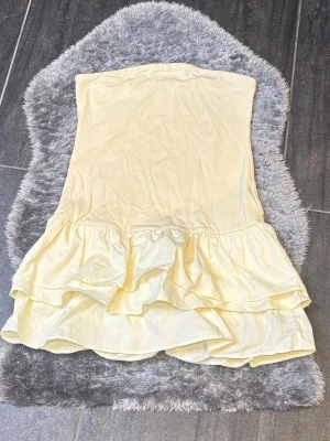 Ljusgul volangklänning Gina Tricot - Söt ljusgul axelbandslös klänning från Gina Tricot med dubbla volanger nedtill. Klänningen har en kort, figurnära passform och är perfekt för sommarens alla tillfällen. Tillverkad i mjukt material som känns skönt mot huden.