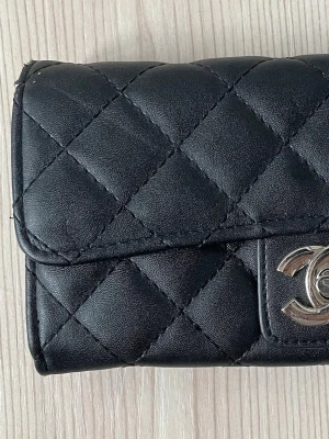 Svart quiltad plånbok  - Säljer en svart quiltad plånbok från Ch i äkta läder. Plånboken har klassiskt rutmönster och ett silverfärgat C..-spänne framtill. Perfekt storlek för kort och kontanter, och ger en lyxig känsla till din vardag.