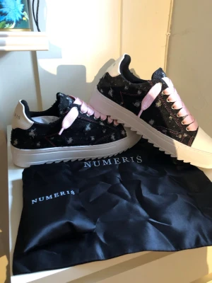 Numeris sneakers med rosa snören - Snygga sneakers från Numeris med svart ovandel i tyg med vita stänk och rosa skosnören. Skorna har vit, räfflad platåsula och vit tå. Märkeslogga på plösen och hälpartiet. Perfekta för dig som vill sticka ut med coola detaljer.