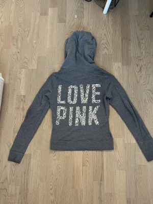 Grå hoodie från PINK med paljetter - Snygg grå hoodie från PINK by Victoria's Secret med dragkedja och huva. På ryggen står det 'LOVE PINK' i stora bokstäver med glittriga paljetter och framtill finns en liten paljettdekorerad hund. Perfekt för dig som gillar en chill och trendig stil. Tröjan har ett litet hål vid vänstra armen, skriv om ni har frågor om det❤️ pris kan diskuteras
