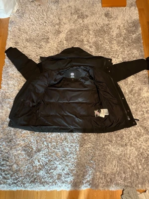 Canada Goose wyndham - Säljer en svart dunjacka från Canada Goose med huva och dragkedja samt knappar framtill. Jackan har flera fickor och ribbade muddar vid ärmsluten. Perfekt för kalla vinterdagar och riktigt varm tack vare dunfyllningen. (Storlek - xs men sitter som S)