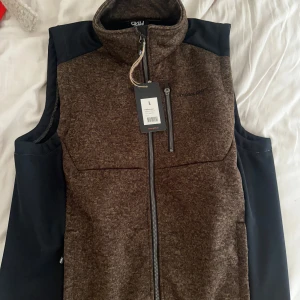 Brun ullväst från P4H  - Snygg brun väst från P4H i ullmix med svarta detaljer på axlar och sidor. Hög krage, hel dragkedja framtill och diskret logga på bröstet. Perfekt lager för kyliga dagar och outdoor-äventyr eller jakt 