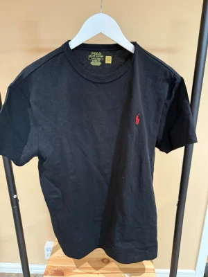 Svart t-shirt från Polo Ralph Lauren - Klassisk svart t-shirt från Polo Ralph Lauren med röd broderad logga på bröstet. T-shirten har rund halsringning och korta ärmar. Tillverkad i mjuk bomull för en bekväm känsla och har en normal passform.