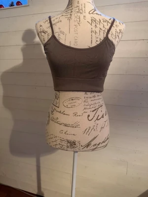 Brun ribbad croppad linnetopp - Säljer en brun croppad linnetopp med tunna axelband och ribbad struktur. Toppen har en enkel och stilren design som är lätt att matcha med olika outfits. Materialet känns mjukt och stretchigt, perfekt för en avslappnad look.