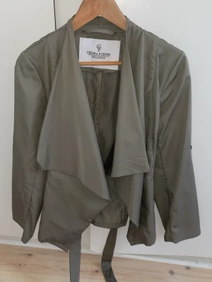 Olivgrön kappa från Chiara Forthi - Stilren olivgrön kappa från Chiara Forti, men köpt från Bubbleroom. Kappan har ett knytband i midjan och långa ärmar. Perfekt nu till våren🌷Jag säljer den för 250kr men är öppen för prisförslag<3