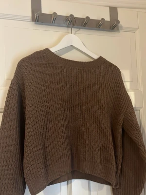 Brun croppad stickad tröja - Mysig brun stickad tröja med croppad passform och grovstickad struktur. Tröjan har rund halsringning och långa ärmar med ribbade muddar. Perfekt för lager på lager och en avslappnad stil.