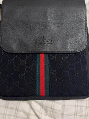 Svart axelväska från Gucci - Snygg svart axelväska från Gucci med klassiskt GG-mönster och ikoniska gröna och röda ränder framtill. Väskan har lock i läder med präglad logga och insida i svart tyg med Gucci-tryck. Perfekt för dig som gillar exklusiva accessoarer.