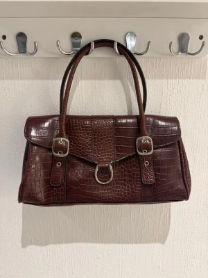 Brun handväska i krokodilimitation - Snygg mörkbrun handväska i krokodilpräglat konstläder med dubbla handtag. Väskan har silverfärgade detaljer, två spännen framtill och en dekorativ ring. Baksidan har en dragkedjeficka för extra förvaring. Perfekt accessoar för en trendig look.