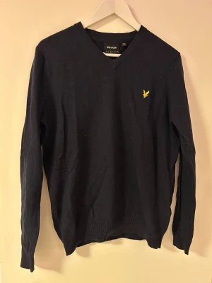 Svart stickad tröja Lyle & Scott - Svart stickad tröja från Lyle & Scott med V-ringning och den klassiska gula logon broderad på bröstet. Tröjan har långa ärmar och ribbade muddar vid ärmslut och nederkant. Perfekt för lager på lager och enkel att matcha.
