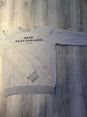 Grå sweatshirt från Peak Performance - Säljer en stilren grå sweatshirt från Peak Performance med svart tryck på bröstet. Tröjan har rund hals, långa ärmar och ribbade muddar. Perfekt för en avslappnad look och gjord i mjukt material.