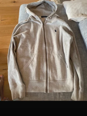 Grå hoodie från Polo Ralph Lauren - Snygg beige hoodie från Polo Ralph Lauren med dragkedja framtill och två fickor. Klassisk huva och den ikoniska lilla loggan broderad på bröstet. Tillverkad i mjukt bomullsmaterial som känns riktigt skönt mot huden. Perfekt för en chill och avslappnad stil.