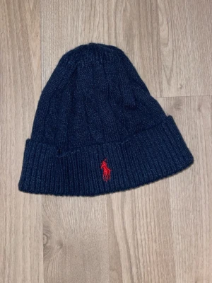 Mörkblå stickad mössa från Polo Ralph Lauren - Säljer en mörkblå stickad mössa från Polo Ralph Lauren med klassisk kabelstickning och uppvikt kant. Mössan har en röd broderad logga framtill och är tillverkad i mjukt material som håller dig varm. Perfekt accessoar för kalla dagar.