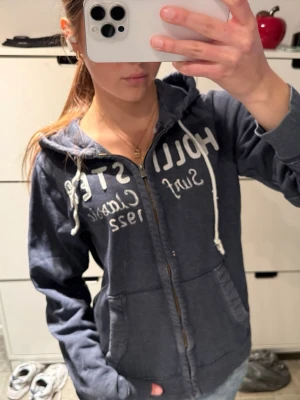Sällsynt Hollister zip up hoodie i en as cool färg - Cool mörkblå färg, hoodie med dragkedja från Hollister.                                                                      Storlek M!                                                                    Stor vit text på bröstet, fickor framtill och vita snören i huvan. Klassisk college-stil och perfekt för chill dagar. Snygg och enkel att matcha till jeans.       Lite slitna snören, lite trögare dragkedja men annars bra skick!