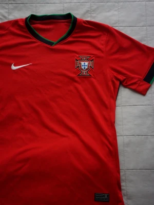 Portugal | Nike fotbollströja  - Snygg röd Portugal fotbollströja från Nike med broderad landslagsemblem på bröstet och vit Nike-logga. Tröjan har gröna detaljer vid kragen och är tillverkad i ett lätt, ventilerande material som passar perfekt till träning eller match.
