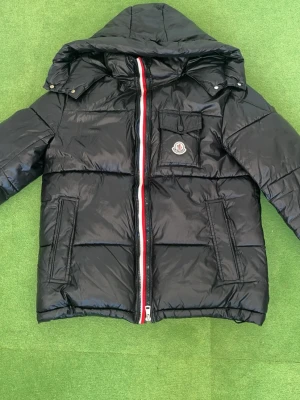 Moncler Jacka - Svart dunjacka från Moncler med glansig finish, huva och coola fickor. Jackan har en rödvit randig dragkedja och Moncler-logga på bröstet. Insidan har det klassiska serietidningsmönstret och flera innerfickor. Perfekt för dig som vill ha en snygg och varm jacka.Montbeliard korta dunjacka för män är en klassisk Moncler-modell. Möts i Stockholm och kan även fraktas. Storlek M sitter mer som S