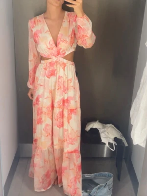 Långklänning - Långklänning med blommigt mönster i rosa och ljusa toner. Klänningen passar perfekt till midsommar men även andra tillfällen! Endast använd en gång, tyvärr belv de ett litet hål men de har sytts igen så de syns ingenting! Storlek 34! 