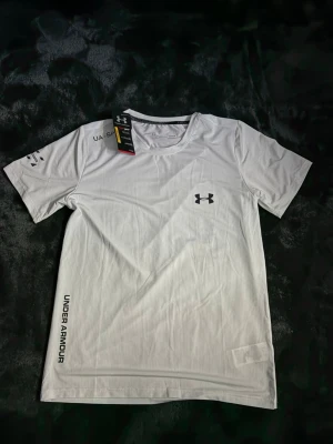 Vit träningströja från Under Armour - Säljer en vit kortärmad träningströja från Under Armour med logga på bröstet och tryck på sidan. Tröjan har rund halsringning och är tillverkad i ett lätt, snabbtorkande material som passar perfekt till träning.