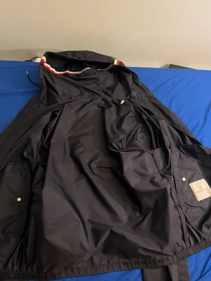 Svart regnjacka från Moncler - Svart regnjacka från Moncler med huva och snygga rödvita detaljer på insidan av huvan. Jackan har praktiska fickor och är gjord i ett vattenavvisande syntetmaterial. Perfekt för regniga dagar och har en clean, modern look.