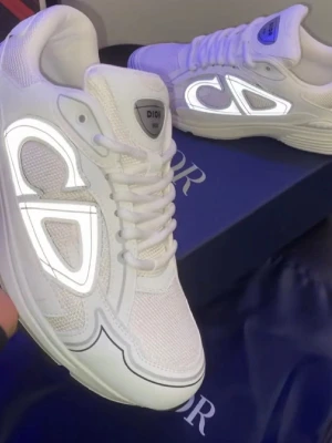 Vita Dior sneakers med reflekterande detaljer - Säljer ett par vita Dior sneakers med stora reflekterande CD-loggor på sidorna. Skorna har en chunky sula, meshpaneler för extra ventilation och vita snören. Perfekt för dig som vill sticka ut med exklusiva och trendiga sneakers.