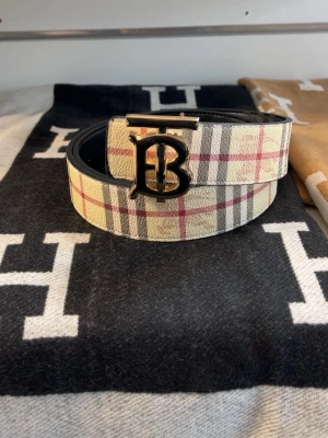 Burberry-bälte reversibel - Snyggt bälte från Burberry med klassiskt rutigt mönster i beige, rött och svart. Bältet har ett stort metallspänne med Burberrys logotyp och är tillverkat i skinn. Perfekt accessoar för att lyfta din outfit.