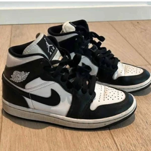 Air Jordan 1 Mid svart/vit sneaker - Snygga Air Jordan 1 Mid sneakers i svart och vitt läder med klassisk Nike swoosh på sidan och Jordan Wings-logga vid ankeln. Skorna har svart snörning, perforerad tåbox och vit mellansula. Perfekta för dig som gillar streetwear och ikonisk design.