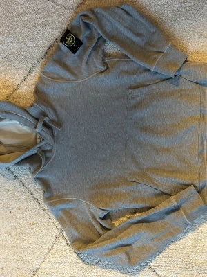 Grå hoodie från Stone Island - Snygg grå hoodie från Stone Island med klassisk logotyp-patch på vänster ärm. Hoodien har huva med dragsko och en stor magficka framtill. Tillverkad i mjukt bomullsmaterial som är skönt mot huden. Perfekt för en avslappnad och trendig stil. Denna tröja är använd ett par gånger för några år sedan, inte använd på ett bra tag. 