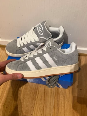 Adidas Campus 00s  - Snygga Adidas Campus sneakers i grå mocka med klassiska vita ränder på sidorna och vit sula. Skorna har lågt skaft och vit insida, samt Adidas-logga på plösen. Perfekta för dig som gillar retrostil och streetwear.