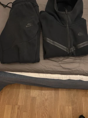 Svart Nike hoodie med dragkedja - Säljer en svart hoodie från Nike med hel dragkedja framtill och diskreta svarta detaljer över bröstet. Hoodien har hög krage, långa ärmar och två fickor. Perfekt för dig som gillar sportig och clean stil, dels byxor till