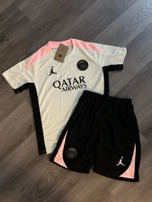 PSG x Jordan träningsset rosa/svart- Exklusiv. S/M junior - Säljer ett PSG x Jordan träningsset med t-shirt och shorts. Tröjan är vit med svarta och rosa detaljer, PSG-logga och Jordan-logga samt Qatar Airways-tryck. Shortsen är svarta med rosa inslag och PSG- samt Jordan-logga. Perfekt för fotbollsträning eller gym. Produkten är S i junior