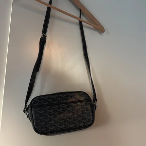 Svart axelväska från  - Snygg svart axelväska från  med klassiskt grafiskt mönster i grått och vitt. Väskan har justerbar rem med vita sömmar, gul insida och dragkedja med metall-detalj. Perfekt storlek för det viktigaste och riktigt trendig look.