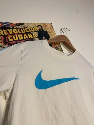 Vit Nike t-shirt med blå swoosh - Vit t-shirt från Nike med stor blå swoosh-logga på bröstet och mindre svarta swoosh-loggor längs ena ärmen. Klassisk rund halsringning och avslappnad passform. Perfekt för dig som gillar sportig och clean stil.