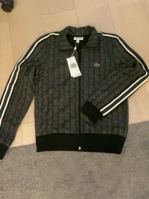 Svart mönstrad ziptröja från Lacoste - Säljer en svart ziptröja från Lacoste med diskret mönster och vita ränder längs ärmarna. Tröjan har dragkedja framtill, ribbade muddar och den klassiska gröna krokodilloggan på bröstet. Perfekt för en sportig och trendig look.