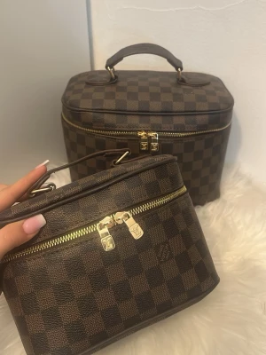 Brun rutmönstrad necessär  - Säljer två klassiska necessärer brunt damier-mönster med guldfärgade dragkedjor och handtag upptill. Perfekta för smink eller småprylar. Lyxig design dem till en riktig statement-accessoar. FAKE 