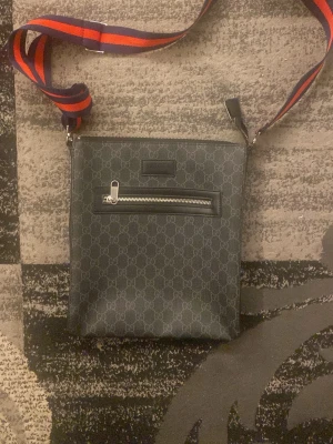 Gucci axelväska med monogram - Snygg svart axelväska från Gucci med klassiskt GG-monogram över hela väskan. Väskan har en framficka med dragkedja och justerbar axelrem i rött och blått. Tillverkad i slitstarkt material med exklusiv känsla och stilren design.