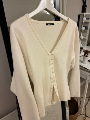 Krämvit kofta från Gina Tricot - Stilren krämvit kofta från Gina Tricot med v-ringning och dekorativa tygklädda knappar framtill. Koftan har långa ärmar och är gjord i ett mjukt, tunt stickat material. Perfekt att matcha med jeans eller kjol för en clean look.