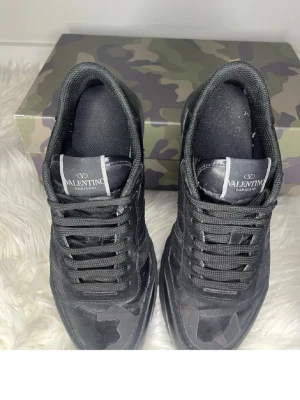 Svarta Valentino Garavani sneakers - Säljer ett par svarta sneakers från Valentino Garavani med coola kamouflagedetaljer och robust sula. Skorna har en mix av läder och textil, samt svarta skosnören och logga på plösen. Perfekta för dig som gillar streetstyle och exklusiva märken. OBS ingen box medföljs 