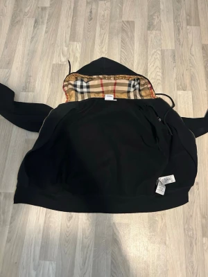 Svart hoodie med Burberry-mönstrad huva - Snygg svart hoodie från Burberry med klassiskt rutigt foder i huvan. Hoodien har dragkedja, långa ärmar och en rymlig passform. Perfekt för dig som vill ha en stilren men ändå unik look. Materialet känns mjukt och bekvämt.